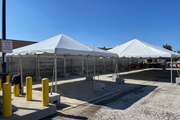 20’x20’ Frame Tent with a solid white roof, no walls, and ballasted with concrete blocks, alongside a 30’x60’ Frame Tent with solid white roof, no walls and ballasted with concrete blocks.